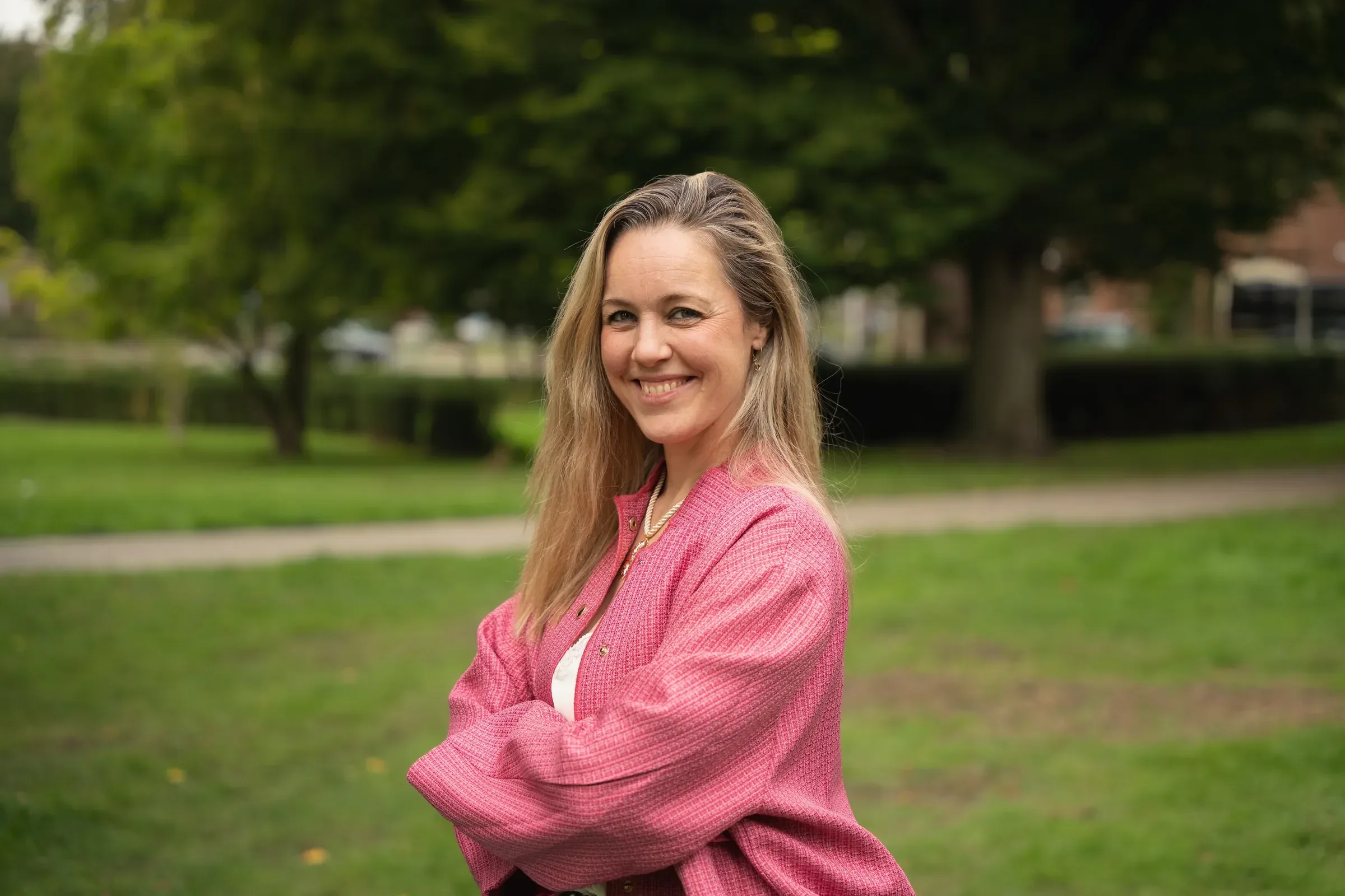 Alies Tax in een roze tweed jasje, glimlachend met gekruiste armen in een groen park.